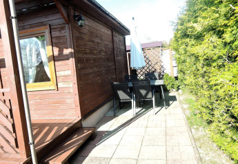 Udendørs terrasse ved en træhytte med bord, stole og parasol, omgivet af grønne buske i solskin.