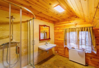 Badkamer in een houten lodge met glazen douche, wastafel, spiegel en raam met gordijnen en radiator.