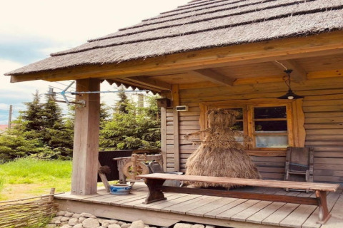 Træhytte med stråtag og halmfigur på verandaen, omgivet af fyrretræer under overskyet himmel.