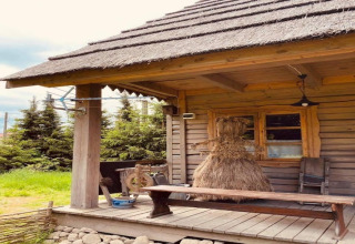 Houten huisje met rieten dak en hooien figuur op het terras, omringd door dennen en een bewolkte lucht.