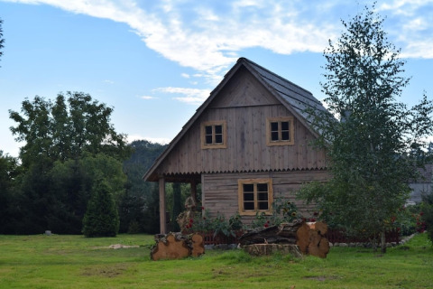 Wooden countryside lodge surrounded by green grass, flowers, and trees beneath a blue sky with clouds.
