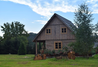 Wooden countryside lodge surrounded by green grass, flowers, and trees beneath a blue sky with clouds.