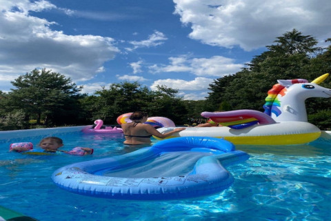 Personer nyder en solrig dag i en pool med børn og farverige oppustelige badelegetøj ved en lodge.