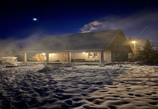 Een sfeervolle blokhut verlicht in de nacht, omgeven door sneeuw en lichte mist onder een donkere sterrenhemel.
