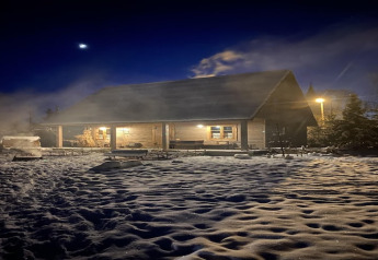 Eine gemütliche Berghütte bei Nacht, umgeben von Schnee und leichtem Dampf unter einem dunklen Himmel.