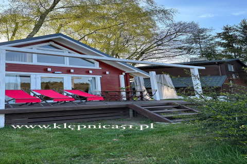 Red beach house with white trim and sun loungers at Lake Klępnica, surrounded by lush greenery.