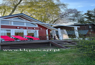 Rood strandhuis met witte accenten en ligstoelen aan het meer Klępnica, omgeven door groen.