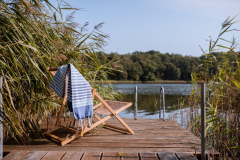 Holzstuhl mit gestreiftem Handtuch auf einem Steg am See Klępnica bei einem Strandhaus in Deutschland.