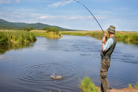 Man fishing by a river near House by the road Skansen in Bełczna, Germany, with scenic green hills.