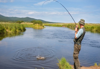 Man fishing by a river near House by the road Skansen in Bełczna, Germany, with scenic green hills.