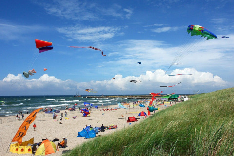 Beach scene with colorful kites flying, people relaxing and playing near House by Lake Klępnica in Germany.