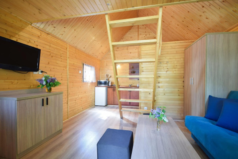 Interieur van een houten vakantiewoning met mezzanine, zetel, kitchenette, tv en bloemen op de tafels.