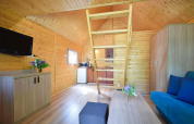 Innenraum eines Holzferienhauses mit Zwischengeschoss, Sofa, Küchenzeile, TV und Blumen auf dem Tisch.