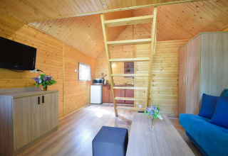 Interior of a wooden holiday cottage with a loft, sofa, kitchenette, TV, and fresh flowers on tables.