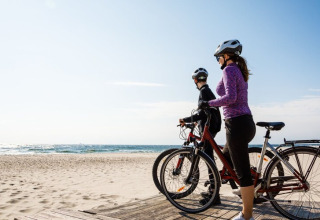 Twee personen op fietsen, met helmen, nemen de zee in zich op bij een glampingverblijf aan het strand.