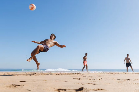 Drie mannen spelen strandvoetbal, iemand maakt een spectaculaire omhaal op het strand met blauwe lucht.