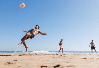 Drie mannen spelen strandvoetbal, één maakt een acrobatische trap op het zand onder een heldere lucht.