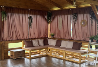 Gezellige overdekte patio met houten banken, bruine gordijnen en veel planten in een vakantiehuis.