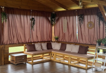 Knusse overdekte veranda met houten palletbanken, bruine gordijnen en planten in vakantiehuis.