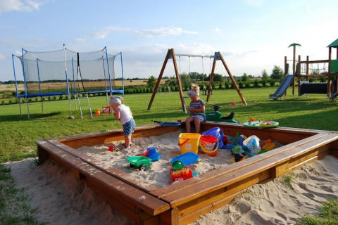 Kinderen spelen in een zandbak met speelgoed op een speelplein met schommels, trampoline en glijbaan.