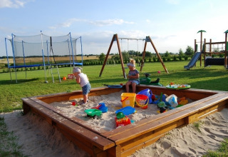 Børn leger i en stor sandkasse ved en legeplads med gynger, trampolin og rutsjebane på en grøn eng.
