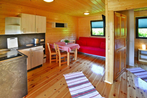 Interieur van een knusse houten blokhut met keuken, eettafel, rode bank en veel daglicht.