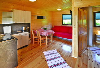 Innenansicht einer gemütlichen Holzhütte mit Küche, Esstisch, rotem Sofa und viel Tageslicht.