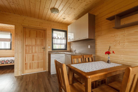 Houten interieur van een vakantiewoning met eettafel, vier stoelen, keuken en slaapkamer op de achtergrond.