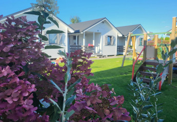 Feriebillede af hvide hytter i Gąski med blomster i forgrunden og legeplads på en grøn græsplæne.