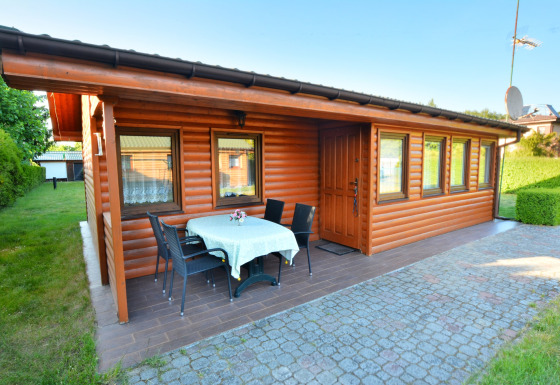Blockhütte mit überdachter Terrasse, Tisch und Stühlen im Garten bei House in Rosnowo, sonniges Wetter.