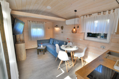Lichte houten cabin-interieur met blauwe bank, eethoek voor twee personen en grote ramen.