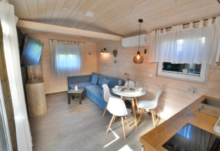 Lichte houten cabin-interieur met blauwe bank, eethoek voor twee personen en grote ramen.
