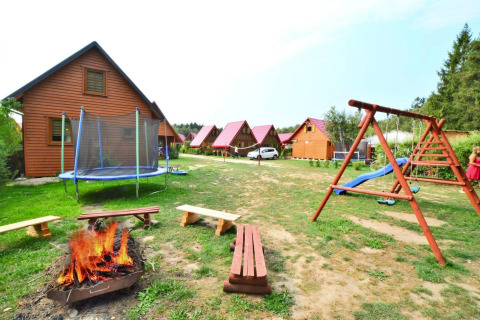 Buitenfoto van een lodge met houten huisjes, speeltuin, trampoline en kampvuur met banken.