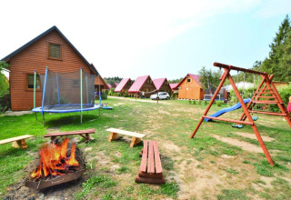 Outdoor scene at a lodge featuring wooden cabins, playground, trampoline, and campfire with benches.