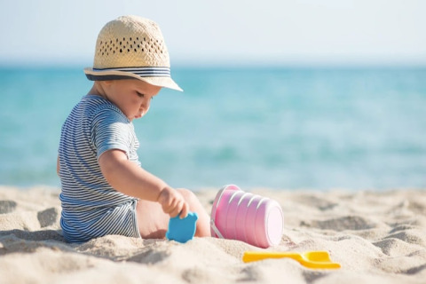 Baby in gestreepte romper met hoedje speelt met strandspeelgoed op het zand bij een glamping aan zee.
