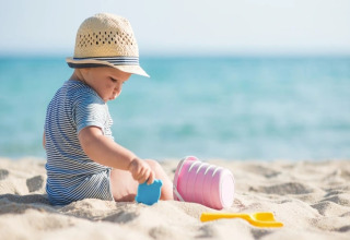 Baby in gestreepte romper met hoedje speelt met strandspeelgoed op het zand bij een glamping aan zee.