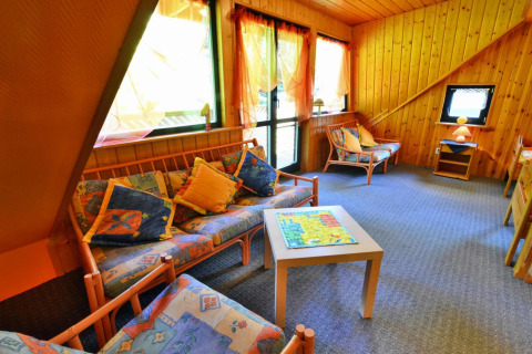 Bright lounge area in a lodge, featuring wooden walls, patterned sofas, and large sunlit windows.