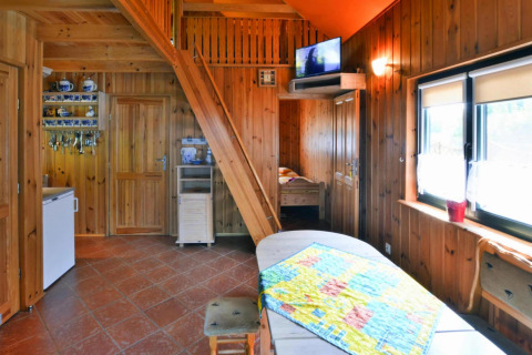 Intérieur de chalet en bois chaleureux avec escalier, TV, coin cuisine et grandes fenêtres lumineuses.