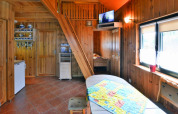 Intérieur de chalet en bois chaleureux avec escalier, TV, coin cuisine et grandes fenêtres lumineuses.