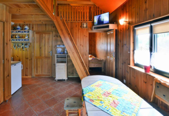 Knus houten lodge-interieur met trap, tv, keukenhoek en grote ramen die veel licht binnenlaten.