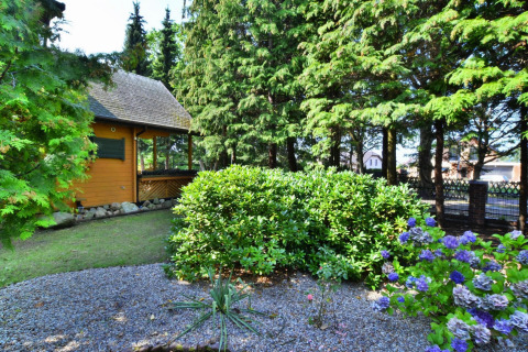Giardino verde con cespugli e fiori davanti a un lodge in legno, circondato da alti pini e luce solare.