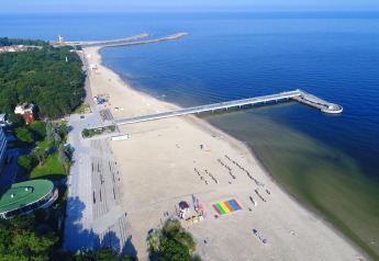 Luchtfoto van een zandstrand en lange pier in Kołobrzeg, Polen, met groene parken aan de kust.