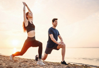 Twee personen doen stretchoefeningen op het strand bij zonsondergang, allebei in sportkleren en relaxed.