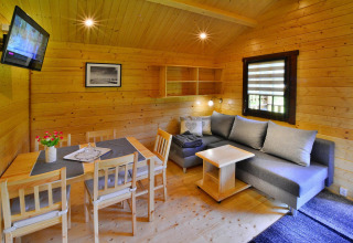 Bright glamping cabin interior with wooden walls, dining table, gray sofa, wall-mounted TV, and window.