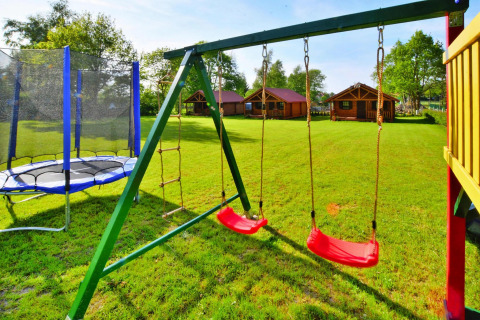 Legeplads med gynger og trampolin foran hyggelige træhytter på glampingplads med grøn græsplæne.