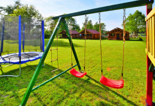 Playground with swings and trampoline in front of cozy wooden cabins at a glamping site on green grass.