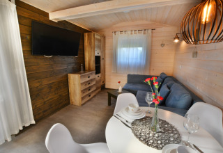 Gezellige woonruimte in een modern houten huisje met blauwe bank, eettafel, tv en natuurlijk licht.