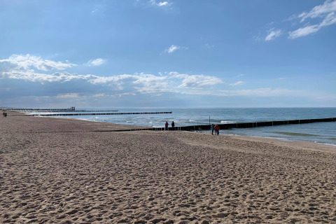 Strandzicht bij Ustronie Morskie met zand, houten steigers en mensen onder een blauwe lucht met wolken.