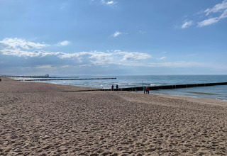 Strandzicht bij Ustronie Morskie met zand, houten steigers en mensen onder een blauwe lucht met wolken.
