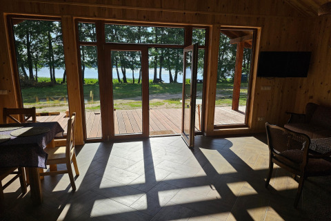 Sunlight pours through large glass doors of a wooden lodge, overlooking trees and a lake outside.